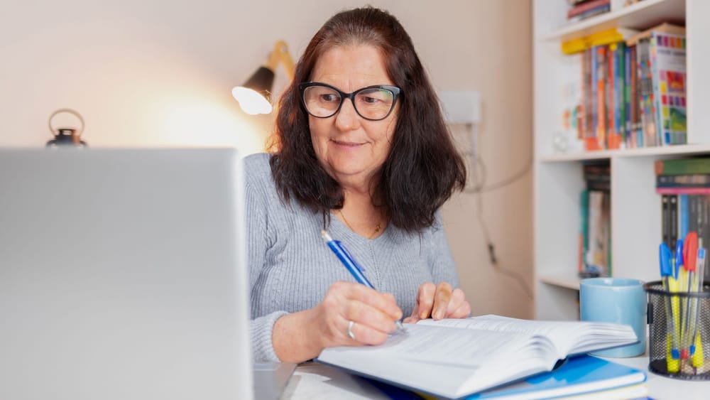 Mulher adulta estudando tranquilamente em casa, escrevendo enquanto usa o notebook